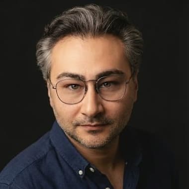 Orkun Camgoz, in a dark blue shirt and round glasses, against a dark studio background.